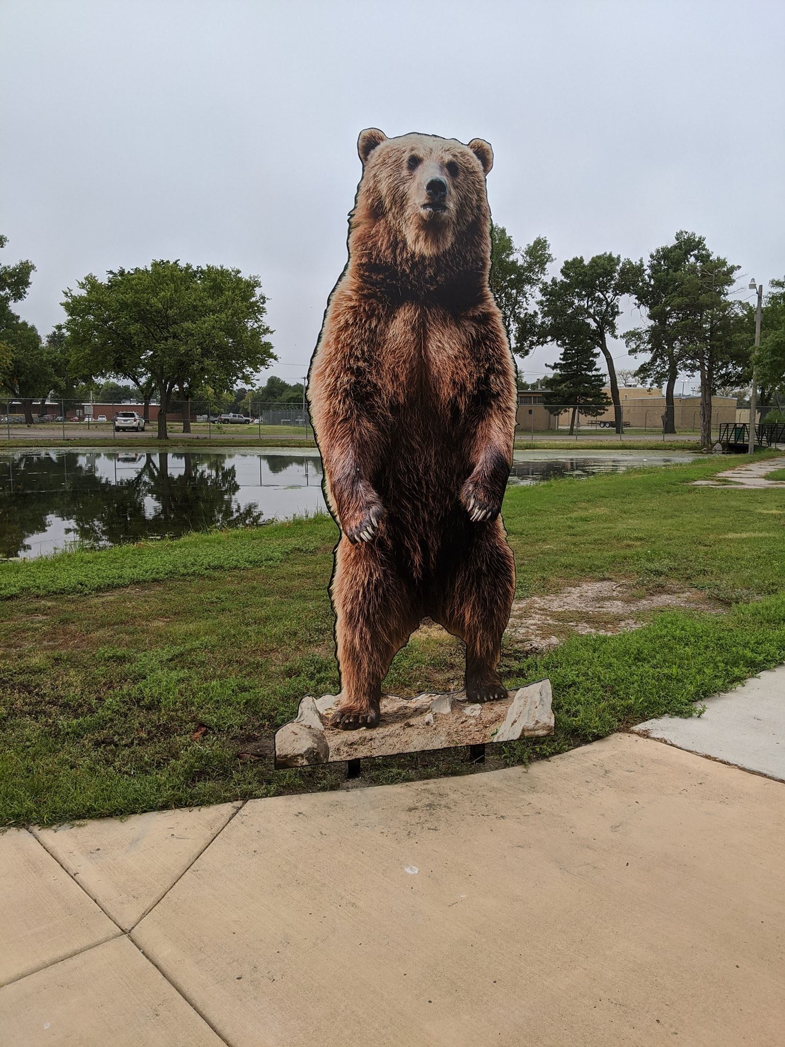 A large bear statue stands prominently in front of a serene pond, surrounded by lush greenery.